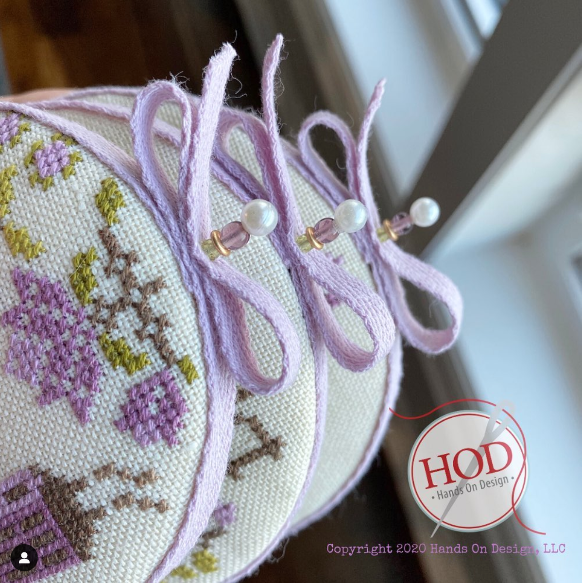 A close-up of a round cross-stitch sampler with lavender twill bows and pearl pins, featuring embroidered floral patterns on a cream fabric background.
Photo by HandsOnDesign.biz [additional_image_link]