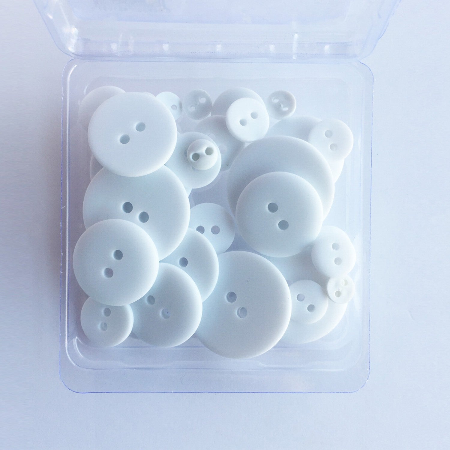 A clear plastic container holding 25 white, matte finish buttons in varying sizes, designed for crafting and sewing projects.
Photo by JustAnotherButtonCompany.com [image_link]