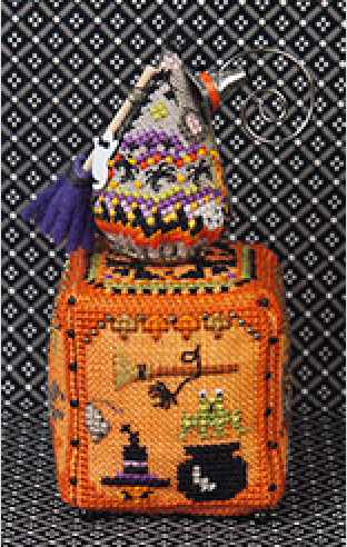 An orange beaded Halloween-themed cube with embroidered motifs including a witch hat, broom, cauldron, bat, and spider, topped with a small mouse beaded ornament and a purple tassel broom, shown against a black-and-white patterned backdrop.
Photo by JustNan.com [additional_image_link]