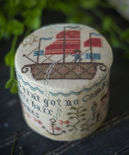 A small cylindrical fabric drum featuring a cross-stitch design of a sailing ship and text on a dark textured surface with blurred green foliage in the background.
Photo by PlumStreetSamplers.com [image_link]